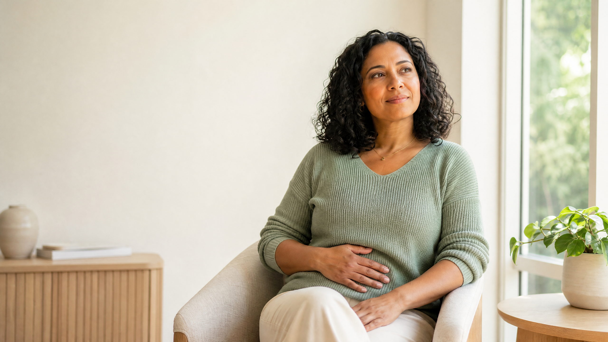 Adult woman sitting in a peaceful, sunlit room with one hand resting gently over her lower abdomen.